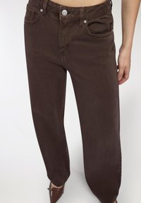 Brązowe dżinsy z wysokim stanem, pięcioma kieszeniami i prostymi nogawkami. Zauważalne srebrne akcenty oraz subtelne detale tekstury.