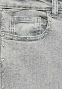 Sive jeans iz džinsa z sprednjo žepom, dekorativnimi kovinskimi bleščicami ob robu in teksturirano površino tkanine. Minimalistični oblikovni detajli.