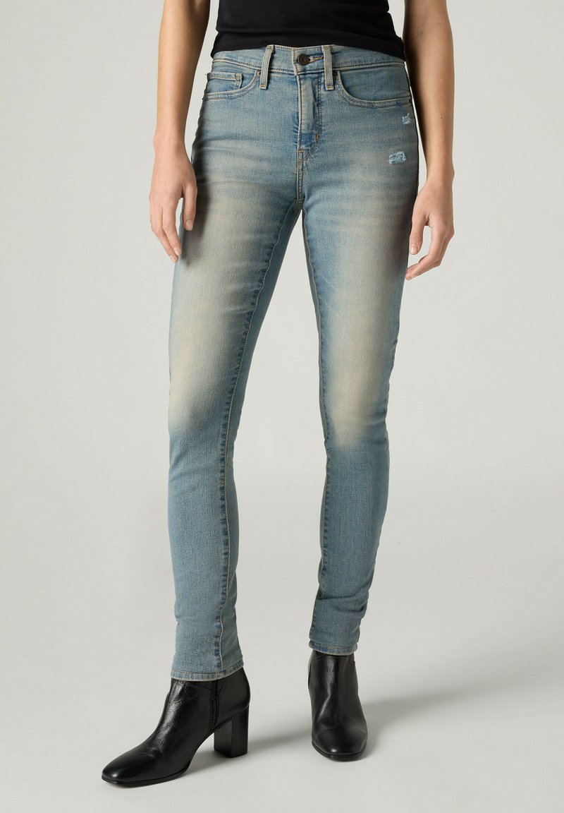 Slim-fit falmede blå jeans med let slid, båret med sorte ankelstøvler med hæl og sort top på neutral baggrund.