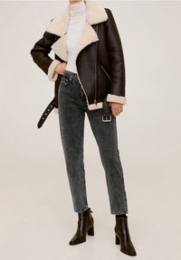 Hnědá kožená bunda s podšívkou z ovčí kožešiny a asymetrickým zipem, nošená přes bílý top. Vytvořeno v kombinaci s šedými džínami s vysokým pasem a černými botami na podpatku.