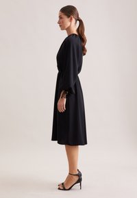 Schwarzes, knielanges Kleid mit V-Ausschnitt, weiten Ärmeln und Taillenkordel. Tragen mit schwarzen, hochhackigen Sandalen mit Knöchelriemen.