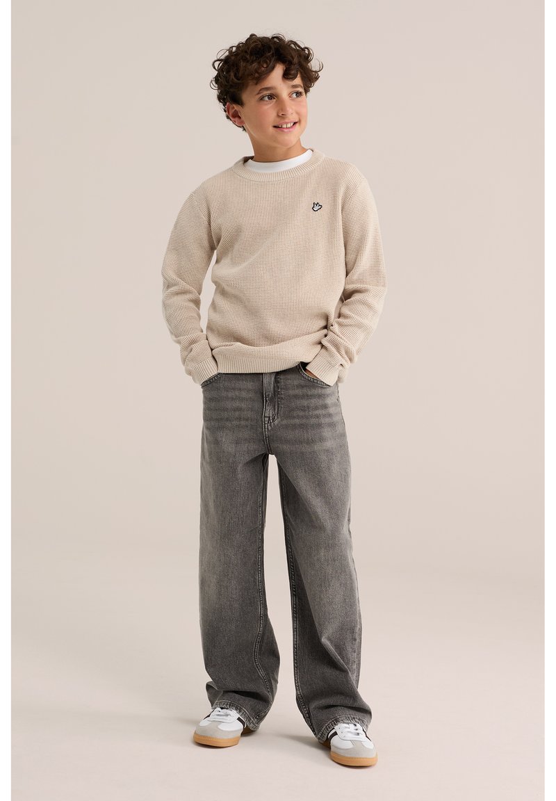 Garçon aux cheveux bouclés portant un pull beige, un jean gris à jambes larges et des baskets blanches, debout les mains dans les poches, regardant vers la droite et souriant.