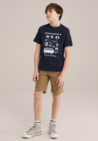 Marinblå bomull T-shirt med vit grafisktryck av sommarartiklar; kombinerad med tan cargo-shorts; grå höga sneakers med vita sulor.
