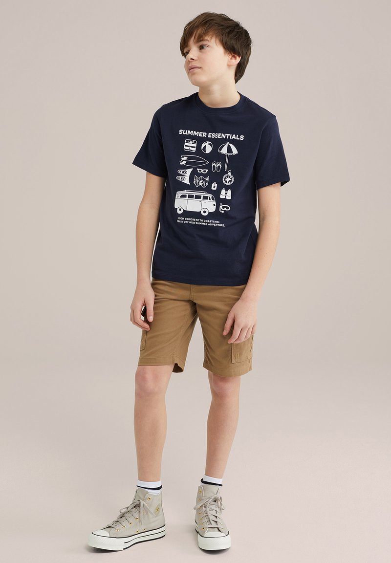 Marinblå bomull T-shirt med vit grafisktryck av sommarartiklar; kombinerad med tan cargo-shorts; grå höga sneakers med vita sulor.