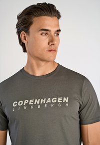 Joven con cabello oscuro que lleva una camiseta gris con el texto "COPENHAGUEN LINDBERGH", mirando hacia su derecha frente a un fondo sencillo.