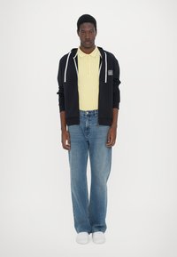 Mornarsko plava zip-up hoodie s bijelim vezicama preko svijetložute polo majice. Svijetloplave hlače širokih nogavica i bijele tenisice upotpunjuju outfit.