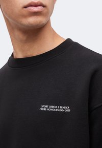 Άνδρας φοράει μαύρο φούτερ με λευκό κείμενο «SPORT LISBOA E BENFICA CLUBS HONOURS 1904-2025» στο στήθος.