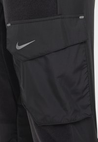 Crne cargo hlače s teksturiranim materijalom i glatkom površinom. Imaju istaknuti sivi Nike logo i veliki džep sa strane.