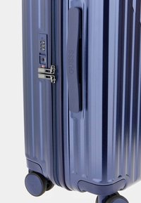 Blaue Hartschalenreisetasche mit vertikalen Rillen, seitlichem Griff, Reißverschluss und Zahlenschloss, ausgestattet mit leichtgängigen Rollen.
