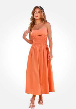 Woman with long wavy hair wearing a sleeveless, orange midi dress with smocked waist and strappy white sandals, posing against a white background.