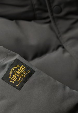Graue Pufferjacke aus einem glatten Stoff. Sie hat ein schwarzes Etikett mit der Aufschrift "SUPERDRY", goldenen Buchstaben und Details in abgerundeter Form.