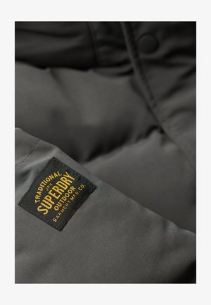 Szary materiał kurtki puchowej o gładkiej fakturze. Posiada czarną metkę z napisem "SUPERDRY", złotymi literami oraz szczegółami w zaokrąglonym kształcie.