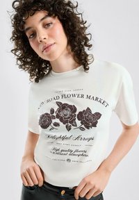 Weißes Baumwoll-T-Shirt mit schwarzem Blumen-Grafikdruck, kurzen Ärmeln und rundem Ausschnitt, gestaltet mit einem dunklen Taillenbund.