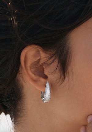 Silberne Tropfenohrringe, verziert mit glitzernden klaren Steinen, mit einer glatten, polierten Oberfläche und einem gebogenen Design, das am Ohr befestigt ist.