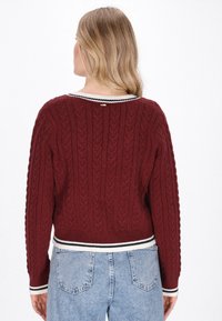 Pulover tricotat cu model în corzi de culoare burgundă, cu o bordură ribbed în dungi alb-negru la tiv și la guler. Include un mic detaliu metalic la gura gâtului din spate.