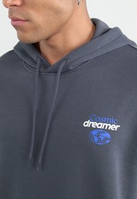 Grey hoodie featuring a "Cosmic dreamer" graphic in blue and white, with a globe design. Soft fabric, adjustable drawstrings, and a relaxed fit.