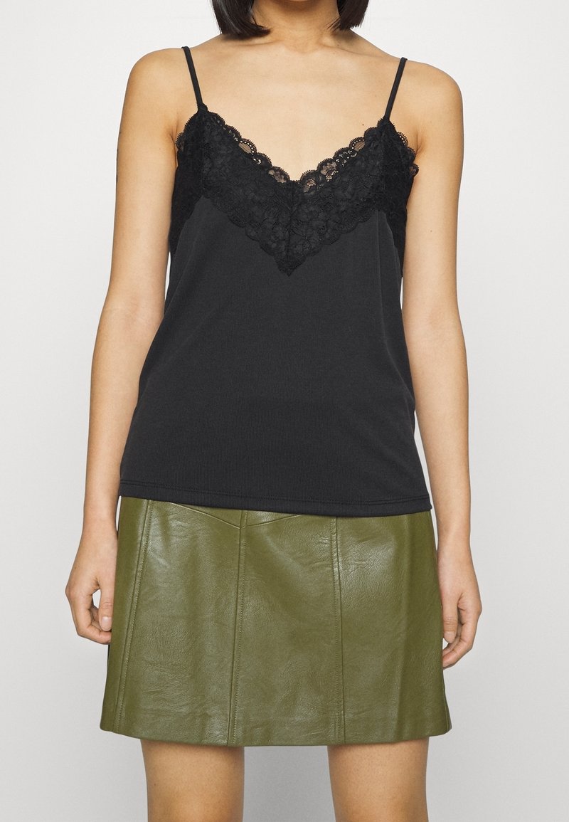 Femme portant un débardeur noir orné de dentelle et une mini-jupe en cuir vert olive, montrée des épaules jusqu'à mi-cuisse.