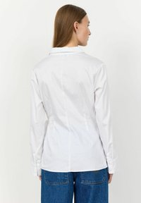 Camicia bianca a maniche lunghe con un design strutturato, caratterizzata da un colletto classico e una texture liscia. Abbinata a jeans larghi blu.