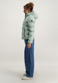 Lichtgroene pufferjack met een capuchon, voorzien van een ritsluiting en zijzakken. G gedragen met wijde spijkerbroeken en witte sneakers.