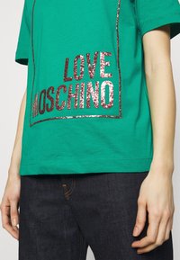 Grön t-shirt med glittrig röd text "LOVE MOSCHINO" som bärs med mörka denimjeans, som visar delar av överkroppen och armarna.