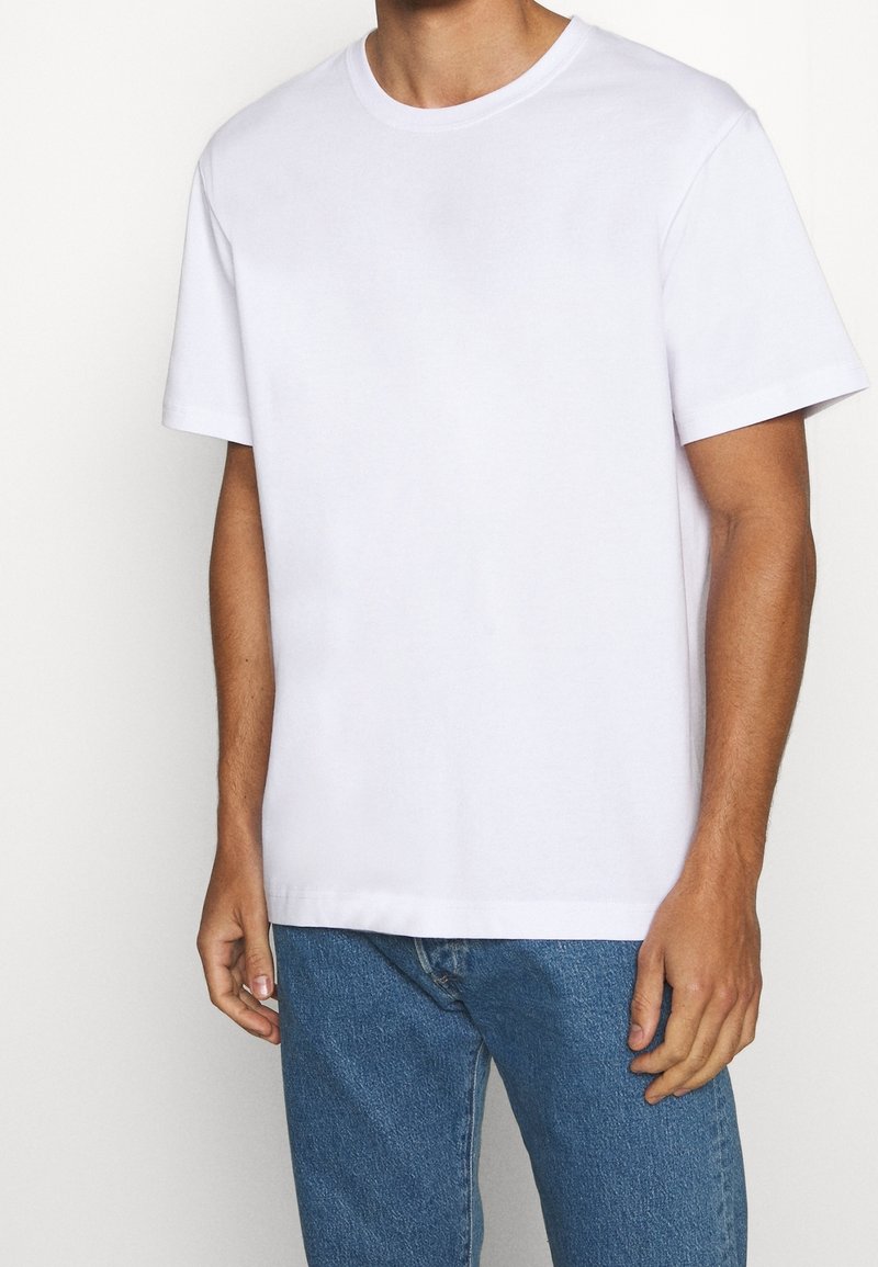 Man som bär en enkel vit t-shirt med korta ärmar och blå jeans, stående mot en vit bakgrund med avslappnad hållning.