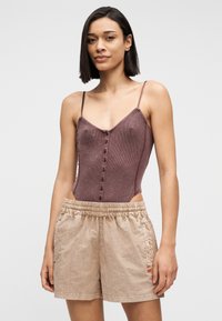 Γυναίκα που φοράει καφέ αμάνικο μπλουζάκι με κουμπιά και άνετα μπεζ σορτς, στέκεται με χαλαρά χέρια μπροστά σε απλό ανοιχτό φόντο.