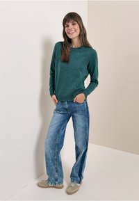 Zielony sweter o fakturowanej powierzchni, luźne niebieskie dżinsy i jasne buty. Sweter ma okrągły dekolt i długie rękawy, co tworzy swobodny fason.