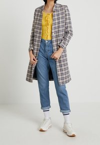 Dlouhý kabát s kostkovaným vzorem v modré, bílé a béžové barvě, přes žluté tričko, doplněný o ohrnuté modré džíny a bílé tenisky s béžovými akcenty.