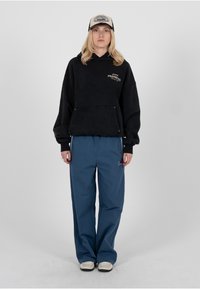 Sweat-shirt à capuche noir avec logo, pantalon cargo bleu et casquette beige. Matière texturée et coupe décontractée ; détail du logo remarquable sur le sweat-shirt.