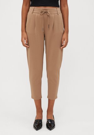 Femme portant un pantalon beige court à cordon et des mocassins noirs vernis à bout pointu, debout devant un fond uni.