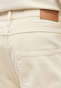 Κοντινή λήψη πίσω τσέπης σε μπεζ τζιν παντελόνι με ραμμένη καφέ δερμάτινη ετικέτα που δείχνει το όνομα της μάρκας "LINDBERGH" στη μέση.