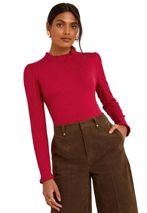 Frau trägt ein eng anliegendes rotes Langarmshirt mit Rüschen an Bündchen und Kragen, kombiniert mit hoch taillierten braunen weiten Hosen und steht mit der Hand in der Tasche.