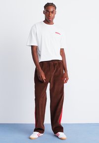 Brown corduroy pants with red side stripes, paired with a white t-shirt featuring a small red logo. Model stands against a neutral background.