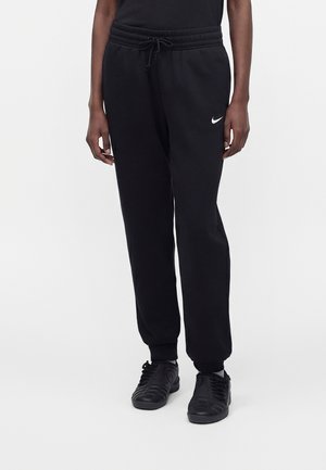 Person iført sorte Nike træningsbukser med snøre i taljen og sorte sneakers, stående mod en ensartet lys baggrund.