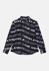 Svart skjorta med långa ärmar och vit text som säger "Versace Jeans Couture" i olika storlekar. Har en knapp-down krage och en rundad nederkant.