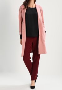 Manteau long rose pâle en tissu texturé, haut noir et pantalon bordeaux, assortis à des talons noirs. Le manteau possède un col cranté et des boutons.