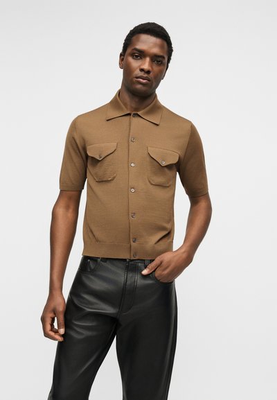 Άνδρας που φορά καφέ κοντομάνικο πουκάμισο με κουμπιά και τσέπες και μαύρο δερμάτινο παντελόνι, στέκεται μπροστά σε απλό ανοιχτό φόντο.