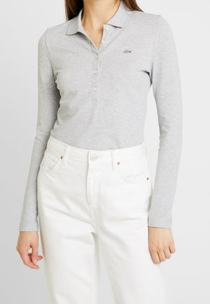 Femme portant un polo à manches longues gris clair avec un petit logo, associé à un pantalon blanc taille haute sur un fond uni.