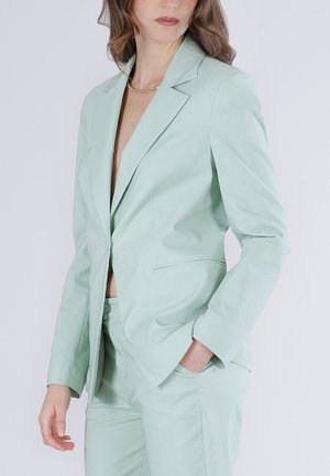 Kobieta ubrana w jasnozielony, dopasowany garnitur z marynarką i pasującymi spodniami, pozująca z jedną ręką w kieszeni na jednolitym tle.