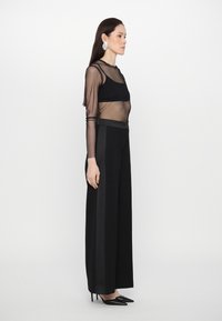 Tenue noire composée d'un haut transparent à manches longues et de pantalons à jambes larges, accompagnée de talons aiguilles noirs et de grandes boucles d'oreilles en argent.