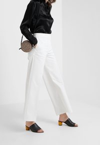 Schwarze Satin-Langarmbluse, weiße hochtaillierte Hose und schwarze Leder-Mules mit Holzabsatz. Runde Handtasche mit Muster.