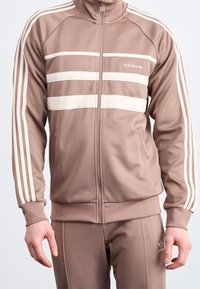 Chaqueta deportiva marrón con tres franjas horizontales blancas en el pecho, cierre de cremallera en la parte delantera y puños acanalados. Fabricada con un tejido suave, cuenta con bolsillos laterales.