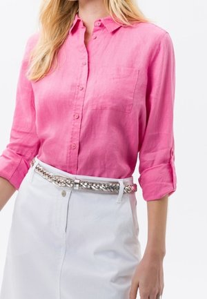 Mujer que lleva una camisa de botones rosa brillante con las mangas arremangadas, metida en una falda blanca con un cinturón metálico trenzado.