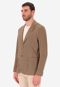 Uomo che indossa un blazer a scacchi marrone e bianco sopra una camicia bianca con pantaloni beige, in piedi contro uno sfondo neutro.