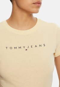 Primer plano de una persona que lleva una camiseta de color beige claro con el texto "TOMMY JEANS" y un pequeño logo rectangular en rojo, blanco y azul en el pecho.