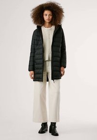 Casaco acolchoado preto com fecho de correr, textura matelassê e capuz. Usado sobre um suéter creme de malha e calças largas bege, combinado com botas pretas.