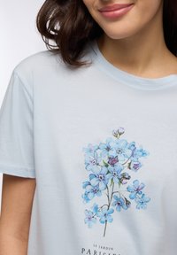 T-shirt di cotone azzurro chiaro con un design floreale in blu e viola, colletto rotondo e maniche corte, stampato con la scritta "Le Jardin Parisien."