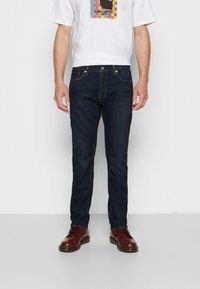 Hombre vistiendo jeans oscuros azules, una camiseta gráfica blanca multicolor y zapatos de cuero marrón con cordones, de pie contra un fondo blanco liso.