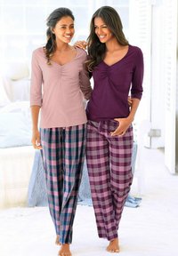 Dos mujeres llevan ropa de estar en casa. Una lleva una camiseta rosa claro con mangas 3/4 y pantalones de cuadros en azul, rosa y verde; la otra lleva una camiseta morada con pantalones de cuadros en morado y rosa.