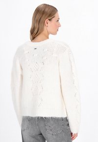 Fehér kötött pulóver laza fazonban, texturált mintázattal és csipke részletekkel. Puha, bolyhos anyag, bordás mandzsettákkal és szegéllyel.